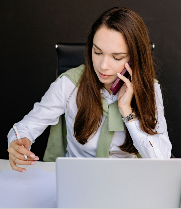 Woman on phone in front of laptop and holding a pen. Navigating the Shift to Value-Based Care: Financial Strategies for a Changing Reimbursement Landscape | Sage